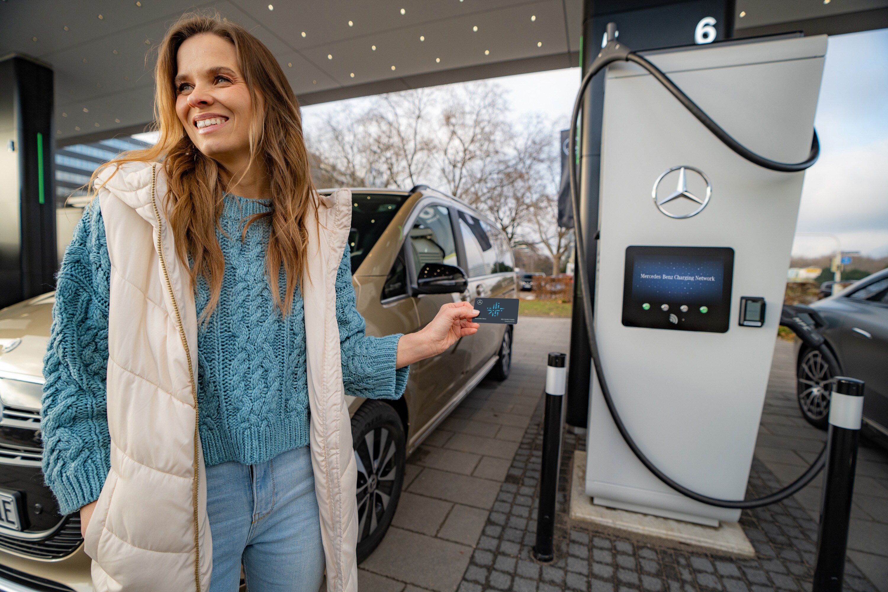 Servicii financiare | Mercedes-Benz Financial Services O femeie este langa o pompa de incarcare electrica. Poarta o vesta lunga gri si tine in mana un card pentru pompa